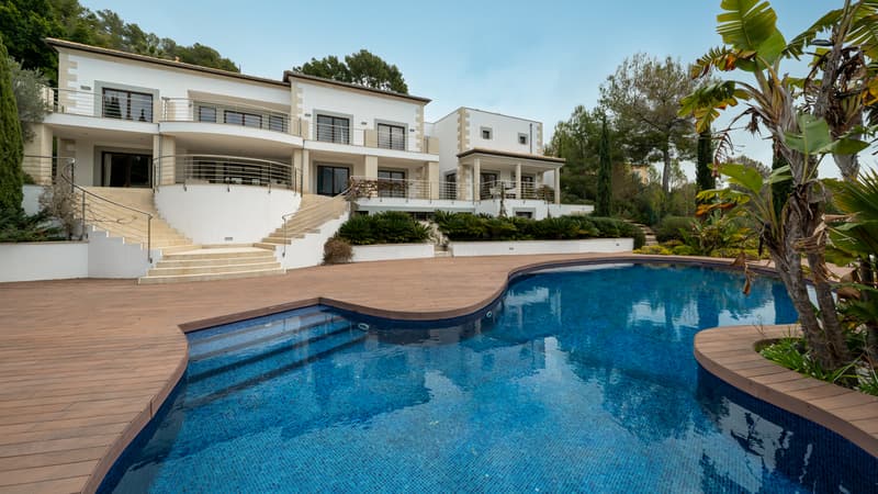 Vue extérieure d'une villa blanche de luxe avec une grande piscine courbe et terrasse en bois entourée de verdure.