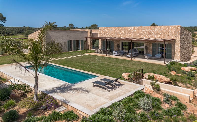 Exterior view of a stone villa with a rectangular swimming pool, lawn, and pergola-covered terrace under a blue sky.
