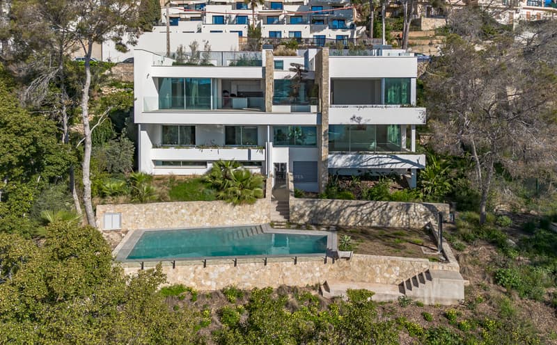 Exterior view of a three-story modern white villa with large windows and a rectangular pool framed by natural stone walls.