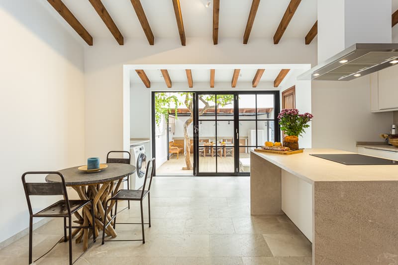 Cuisine lumineuse avec poutres en bois, îlot en pierre et portes vitrées s'ouvrant sur un patio avec un arbre.