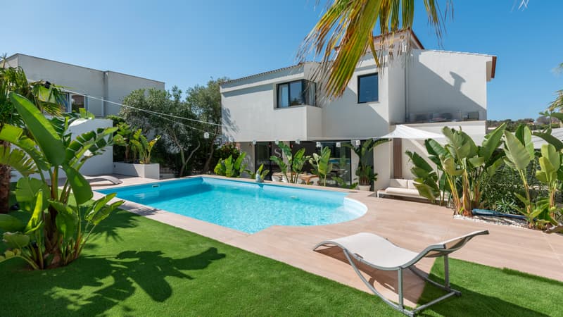 Modern vit villa med blå pool, solsäng, grön gräsmatta och tropiska växter under en klarblå himmel.