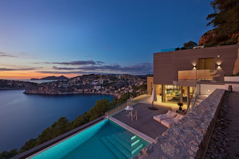 Terrasse d'une villa moderne avec piscine à débordement surplombant une baie au coucher du soleil.