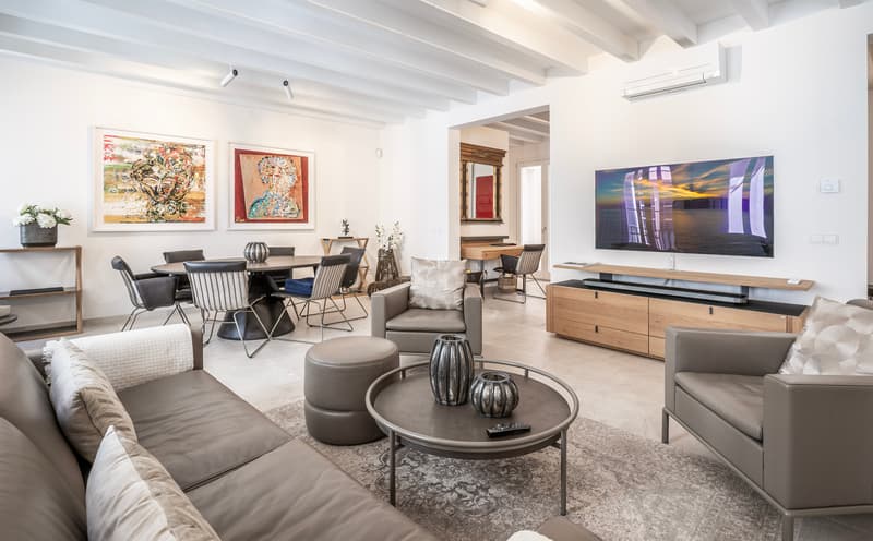 Geräumiges modernes Wohnzimmer mit grauen Ledersofas, Holzbalkendecke, großem Fernseher und angrenzendem Essbereich mit Kunst.