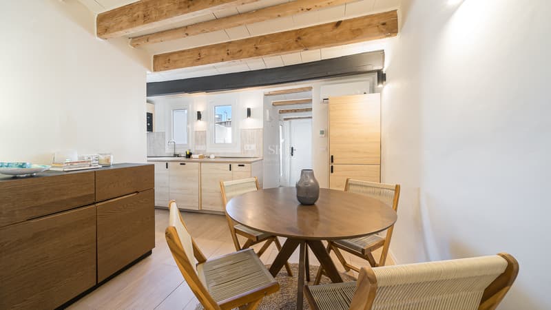Salle à manger avec table ronde en bois, chaises en corde et poutres d'origine menant à une cuisine moderne.
