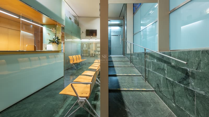 Espace de réception avec sol en marbre vert, chaises en bois, parois en verre dépoli et escalier en acier.