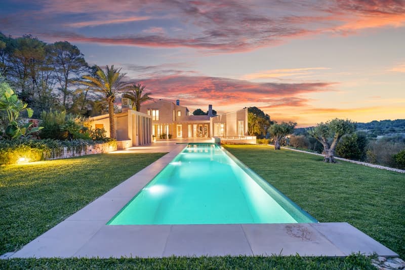 A long, illuminated rectangular pool leading to a modern stone villa under a vibrant pink and orange sunset sky.