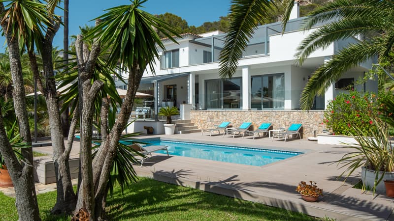 Moderne weiße Villa mit rechteckigem Pool, Sonnenliegen und Palmen unter blauem Himmel.