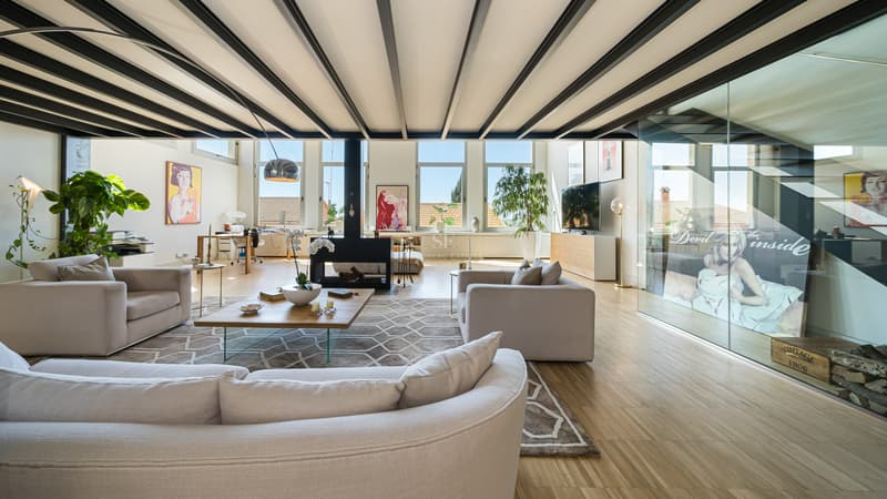 Ljust vardagsrum med svarta stålbalkar, krämfärgade soffor, central öppen spis och stora fönster.