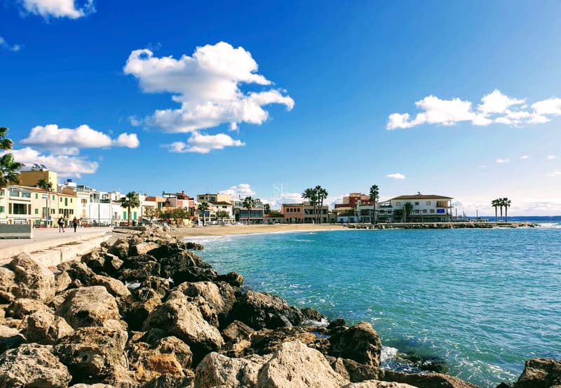 Vue depuis un rivage rocheux vers une plage de sable et des bâtiments côtiers sous un ciel bleu vif.