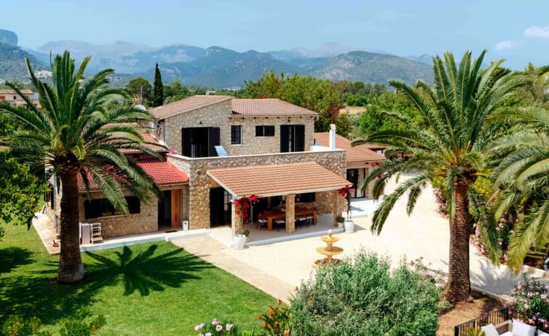 Mediterrane Steinvilla mit Palmen, gepflegtem Rasen und Bergen im Hintergrund unter blauem Himmel.