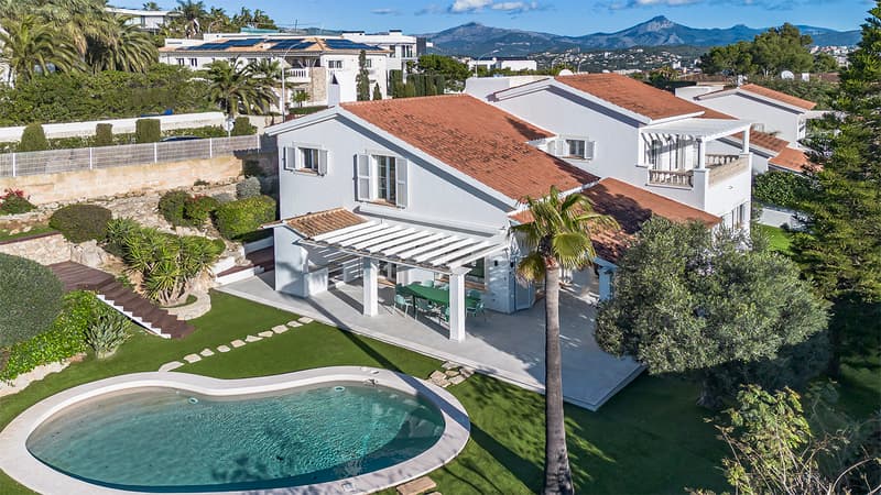 Vista aérea de una villa blanca con tejado de terracota, piscina riñón y jardín con montañas al fondo.