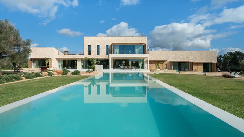 Modern tvåvåningsvilla med en stor rektangulär pool och naturstensfasad under en blå himmel.