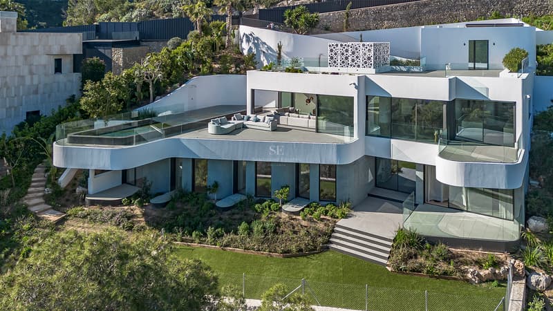 High-angle view of a multi-level white modern villa with an infinity pool, curved terraces, and lush gardens.