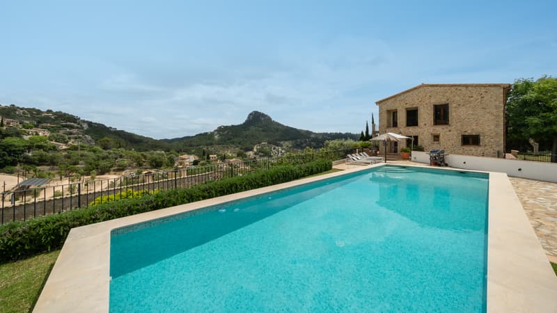 Grande piscine rectangulaire surplombant une vallée verdoyante et des montagnes avec une villa en pierre.