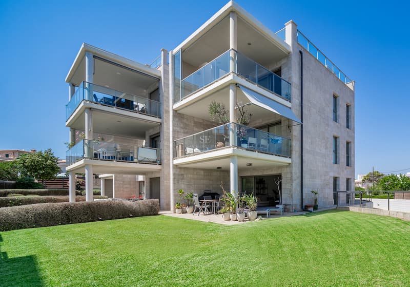 Villa contemporaine de trois étages avec revêtement en pierre, balustrades en verre et pelouse verte sous un ciel bleu.