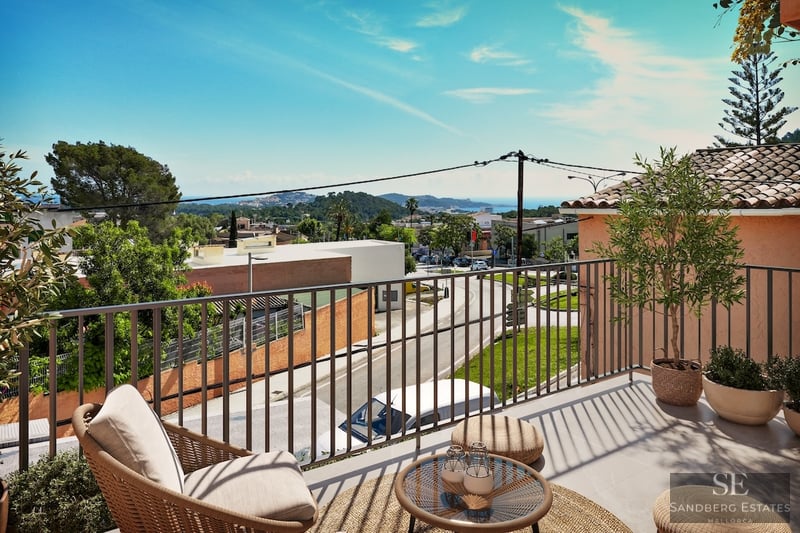 A sunny elevated terrace with a wicker armchair and small table overlooking a coastal town and the sea.