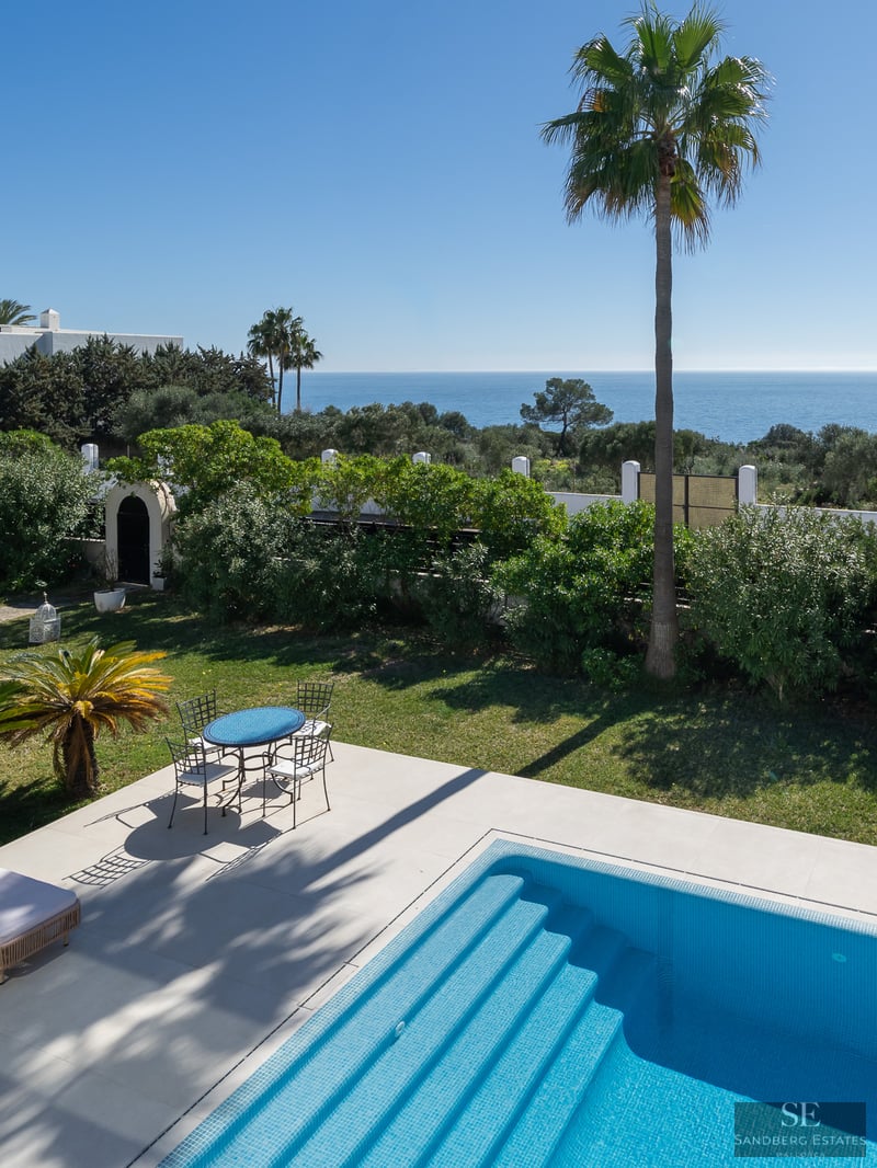 Una piscina azul brillante y terraza con comedor exterior frente a un jardín exuberante y el mar Mediterráneo.
