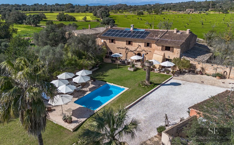 High angle aerial shot of a stone villa with a blue pool, solar panels, and white umbrellas surrounded by green fields.