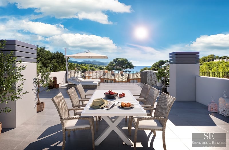Sun-drenched rooftop terrace with white dining set, lounge chairs, and a panoramic view of the sea and coastal hills.