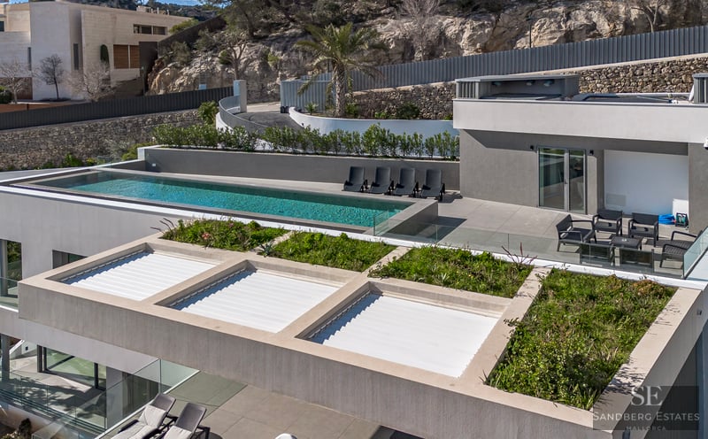 High-angle view of a modern grey villa featuring an infinity pool, sun loungers, and rooftop greenery against a rocky hill.