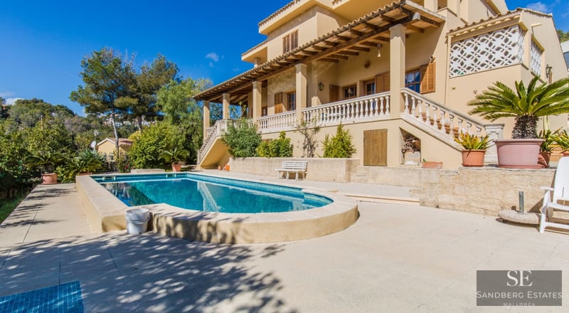 Gran piscina privada frente a una villa mediterránea de color ocre con terraza y colinas verdes al fondo.