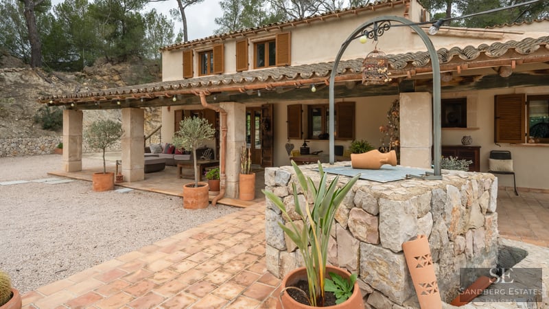 Fachada de casa rústica con porche cubierto, contraventanas de madera y un pozo de piedra en primer plano.