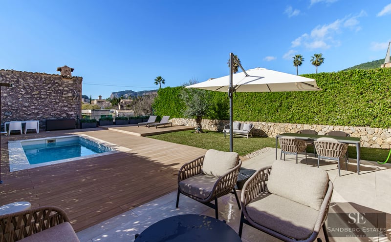 Modern swimming pool with wooden deck, stylish outdoor furniture, and lush green hedges under a clear blue sky.