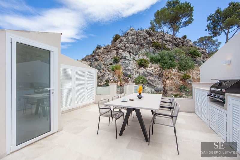 A modern white terrace with a dining table, chairs, and a barbecue area, set against a rocky mountain backdrop.