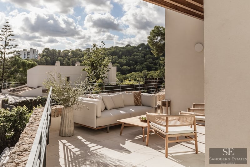 Outdoor terrace with modern sofa, wooden table, and woven chairs overlooking a lush green valley.