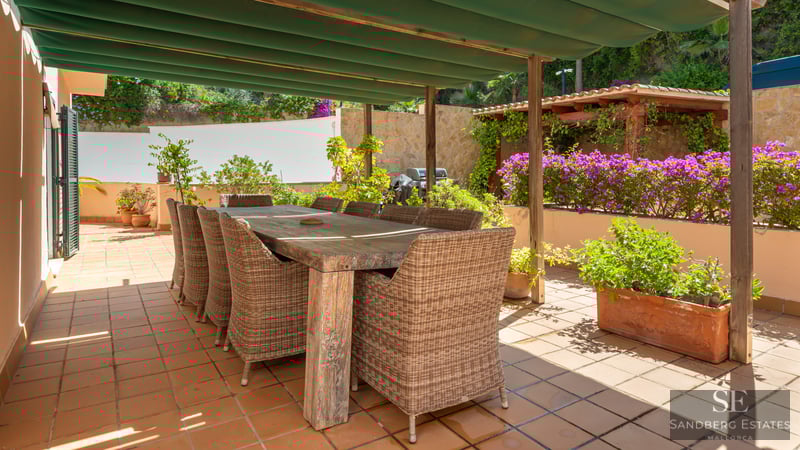 Large wooden dining table with wicker chairs on a sunny terracotta terrace under a green awning.