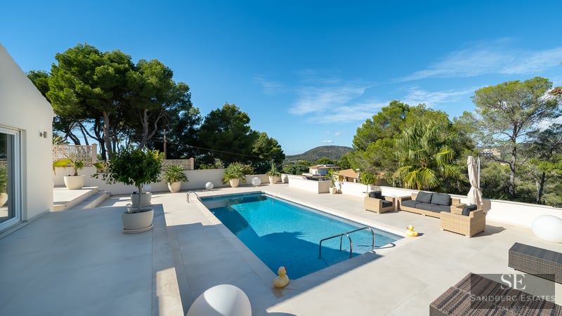 Piscine bleue rectangulaire sur terrasse blanche avec mobilier de jardin et vue sur la forêt méditerranéenne.