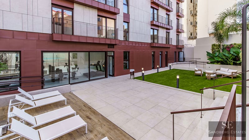 Innenhof mit Steinplatten, Sonnenliegen, Rasen mit Sofas und verglastem Fitnessstudio unter modernem Gebäude.