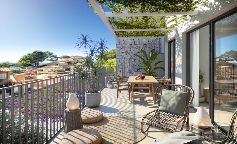 Sunny modern terrace with a wooden dining table, wicker lounge chair, and a plant-covered pergola overlooking a coastal town.