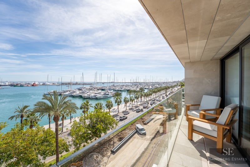 Modern balcony with wooden armchairs and glass railing overlooking a marina filled with yachts and palm trees.