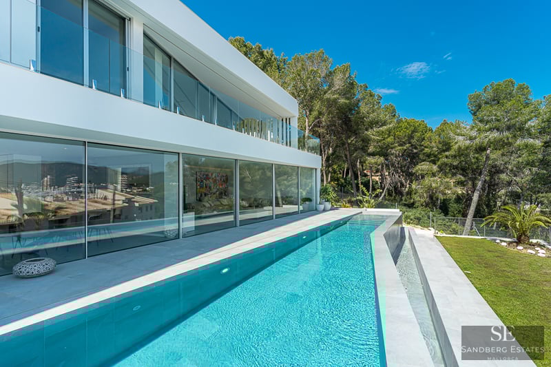 A sleek modern white villa with a long turquoise lap pool, floor-to-ceiling glass windows, and lush green trees.
