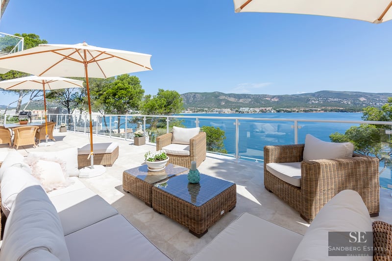 Sonnige Terrasse mit Korbmöbeln, weißen Sonnenschirmen und Glasgeländer mit Blick auf das Meer und die Berge.