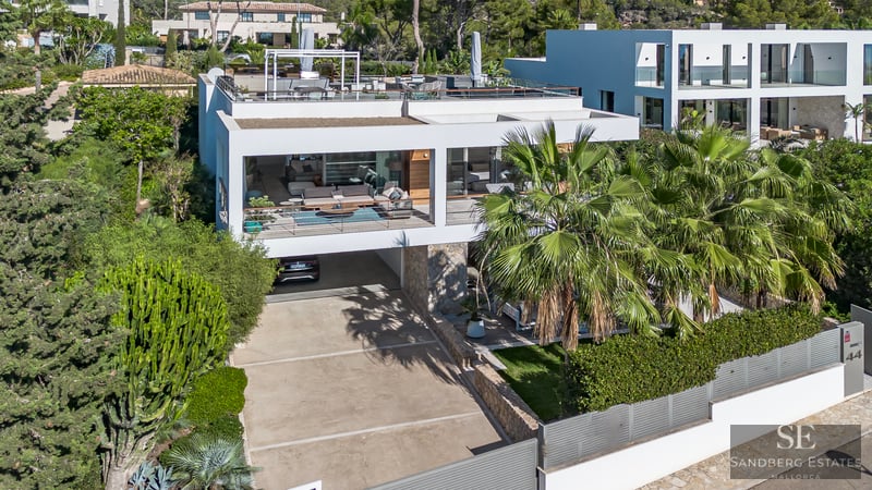 Vue architecturale d'une villa moderne blanche avec terrasses, palmiers et murs en pierre sous une lumière vive.