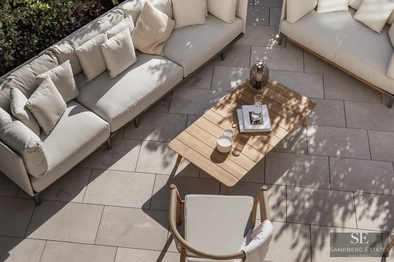 High-angle view of a luxury terrace with beige sofas, a wooden coffee table, and stone paving in dappled sunlight.