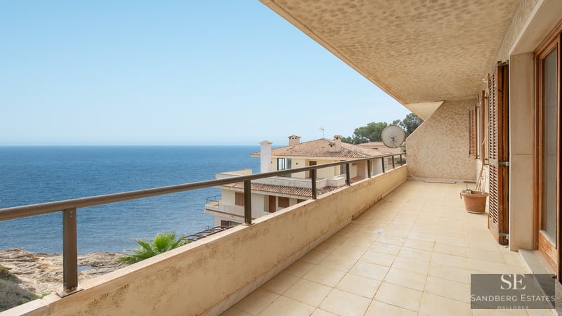 A wide tiled balcony with a metal railing overlooking the blue ocean and nearby Mediterranean buildings.
