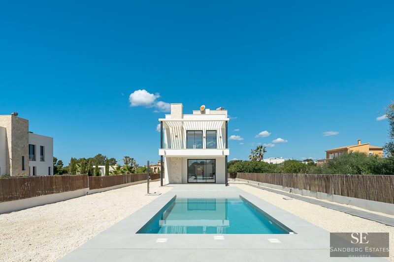A rectangular blue swimming pool leading to a modern white two-story villa under a clear blue sky.
