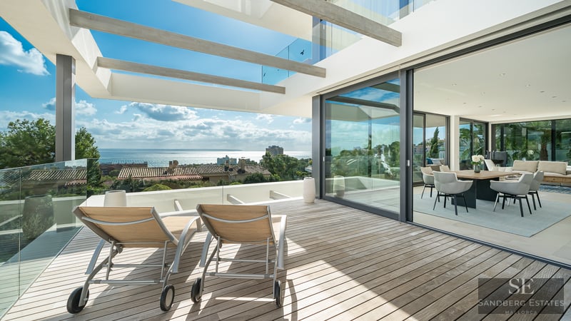 Sun loungers on a wooden terrace with glass railings overlooking the sea, adjacent to an open-plan dining room.