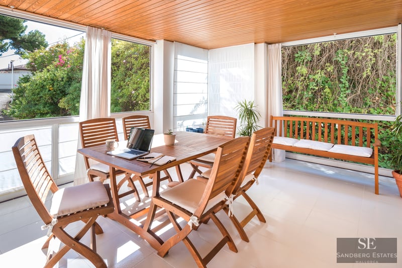 Ein sonnendurchfluteter Terrassenraum mit Esstisch aus Holz, Stühlen und Bank, mit Blick auf das üppige Grün des Gartens.