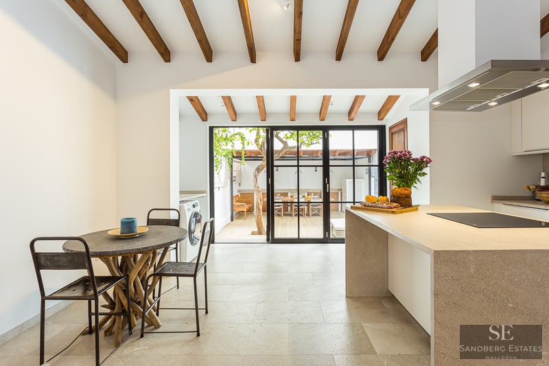 Bright kitchen with wood ceiling beams, stone island, and glass doors opening to a sunny outdoor patio with a tree.