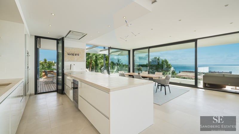 Bright modern white kitchen with large island, dining area, and floor-to-ceiling glass walls overlooking the ocean.
