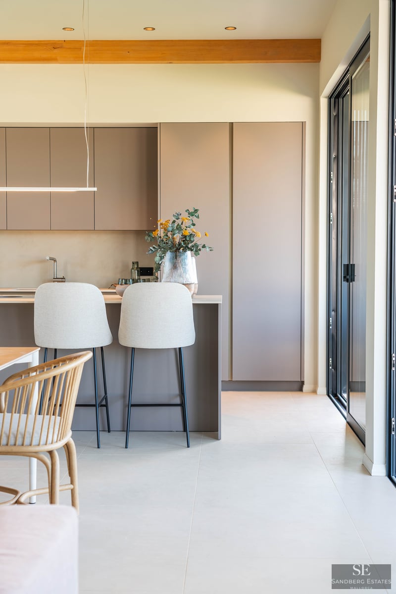 A modern kitchen featuring matte taupe cabinets, an island with two light-colored bar stools, and a wooden ceiling beam.