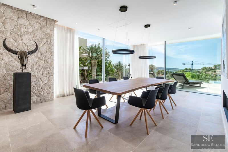 A modern dining room featuring a large wooden table, black chairs, a stone wall with a skull sculpture, and large windows.