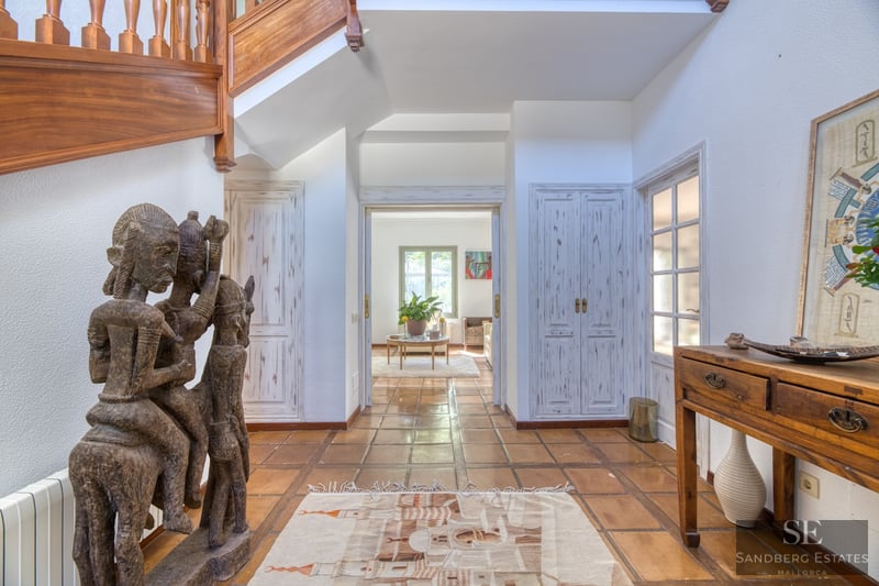 Amplio pasillo con suelos de terracota, una gran estatua tribal de madera y una escalera de madera hacia arriba.