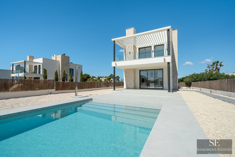 Clear blue swimming pool with steps, next to a modern two-story white and stone villa under a bright blue sky.