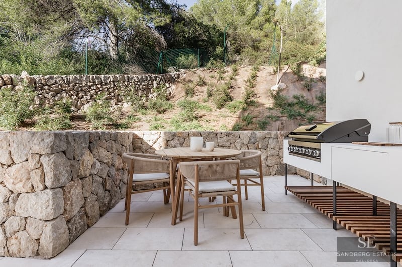 Modern Terrace with Outdoor Kitchen and Dining Outdoor terrace featuring a wooden dining set, stone walls, and a modern built-in grill area against a backdrop of trees.