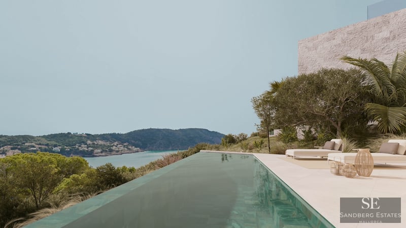 Infinity swimming pool on a stone terrace overlooking a turquoise bay and green hills under a clear sky.